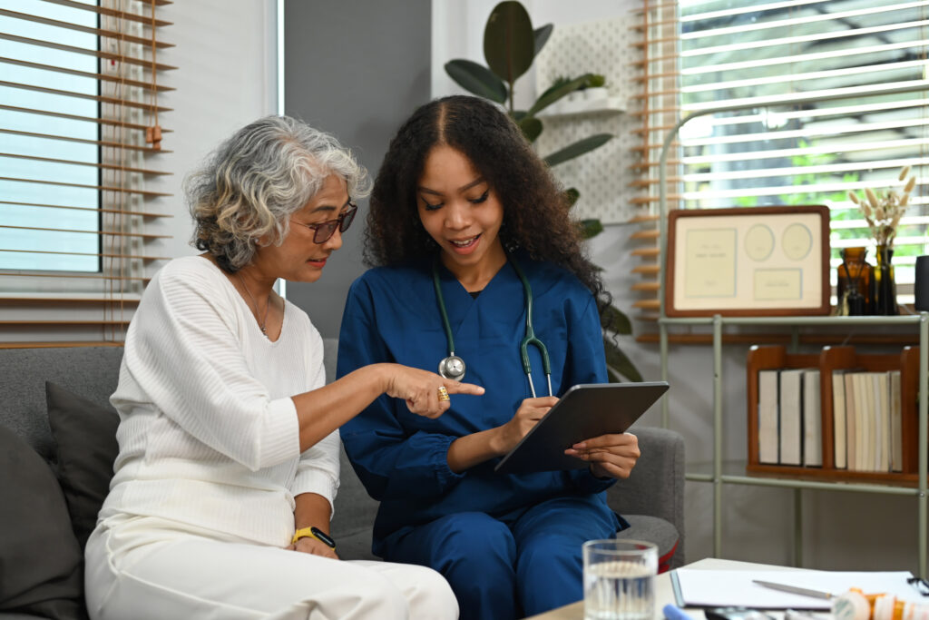 Practitioner nurse showing medical report to senior woman on digital tablet during home care visit.